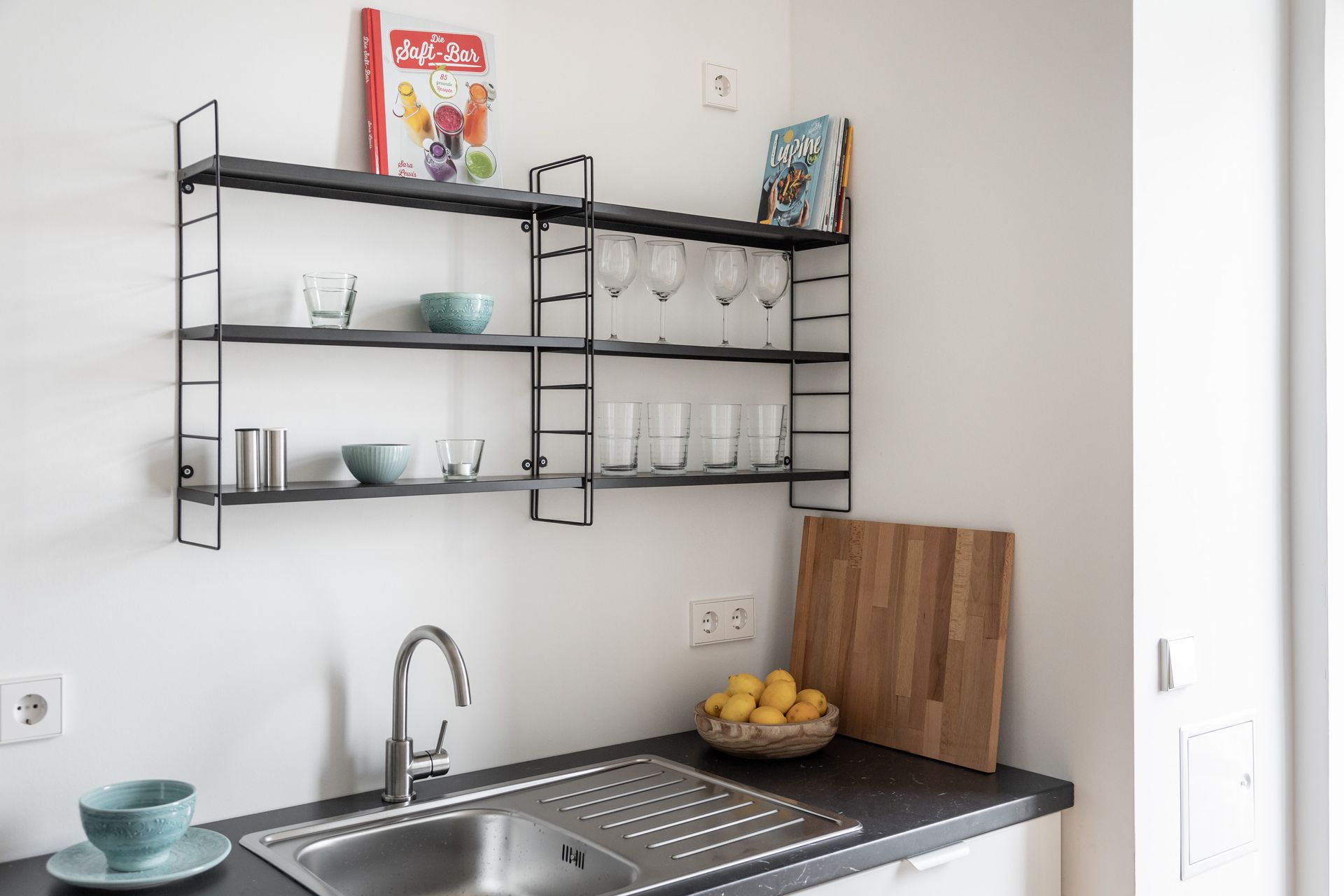 A kitchen with a sink , shelves , glasses , bowls and a cutting board.