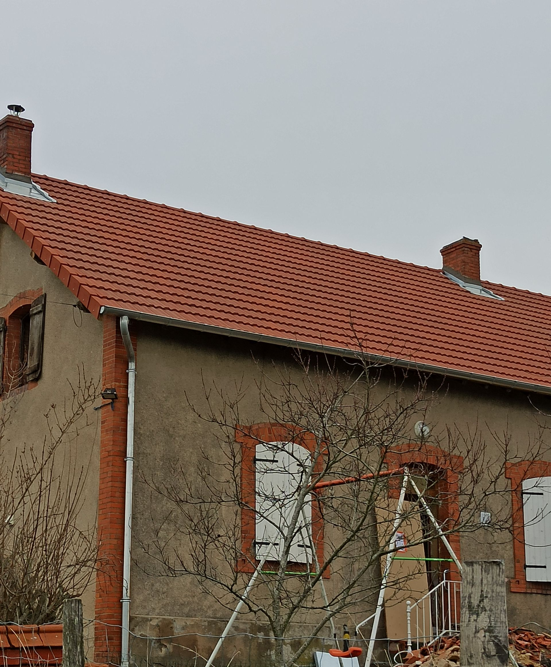 Maison en stuc patiné par le temps, avec un toit de tuiles rouges et des fenêtres à cadre blanc, sous un ciel gris.