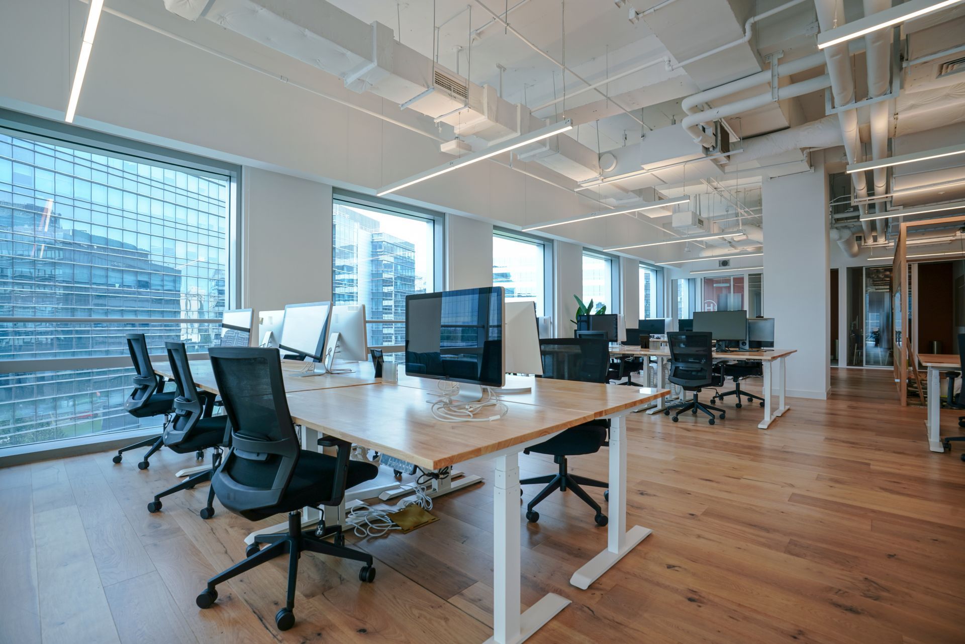 Oficina moderna y luminosa con escritorios, sillas, computadoras y grandes ventanales con vista al paisaje urbano.