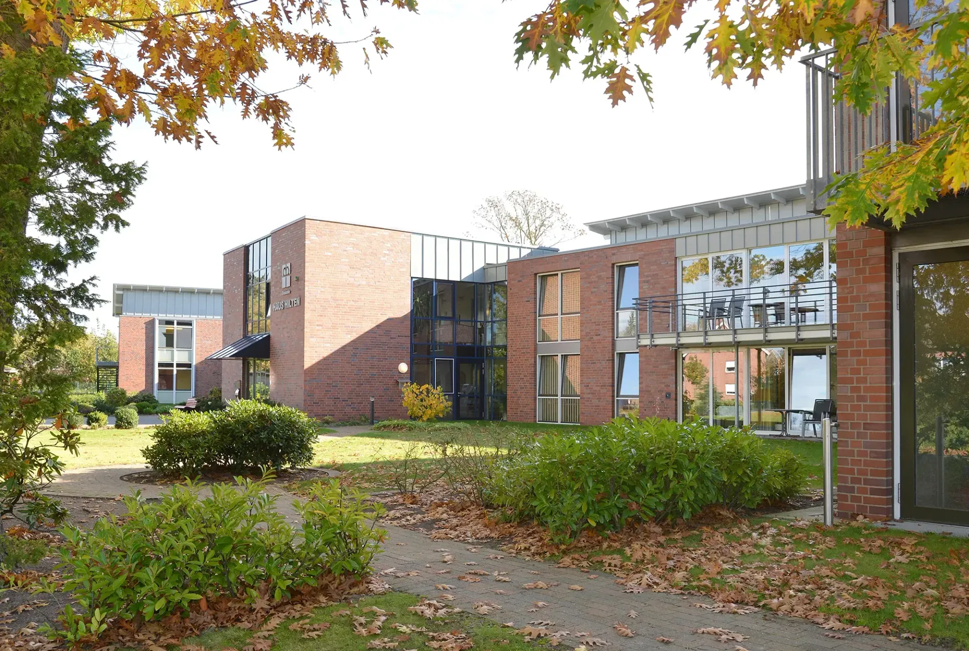 Ein großes Backsteingebäude mit vielen Fenstern ist von Bäumen und Büschen umgeben.