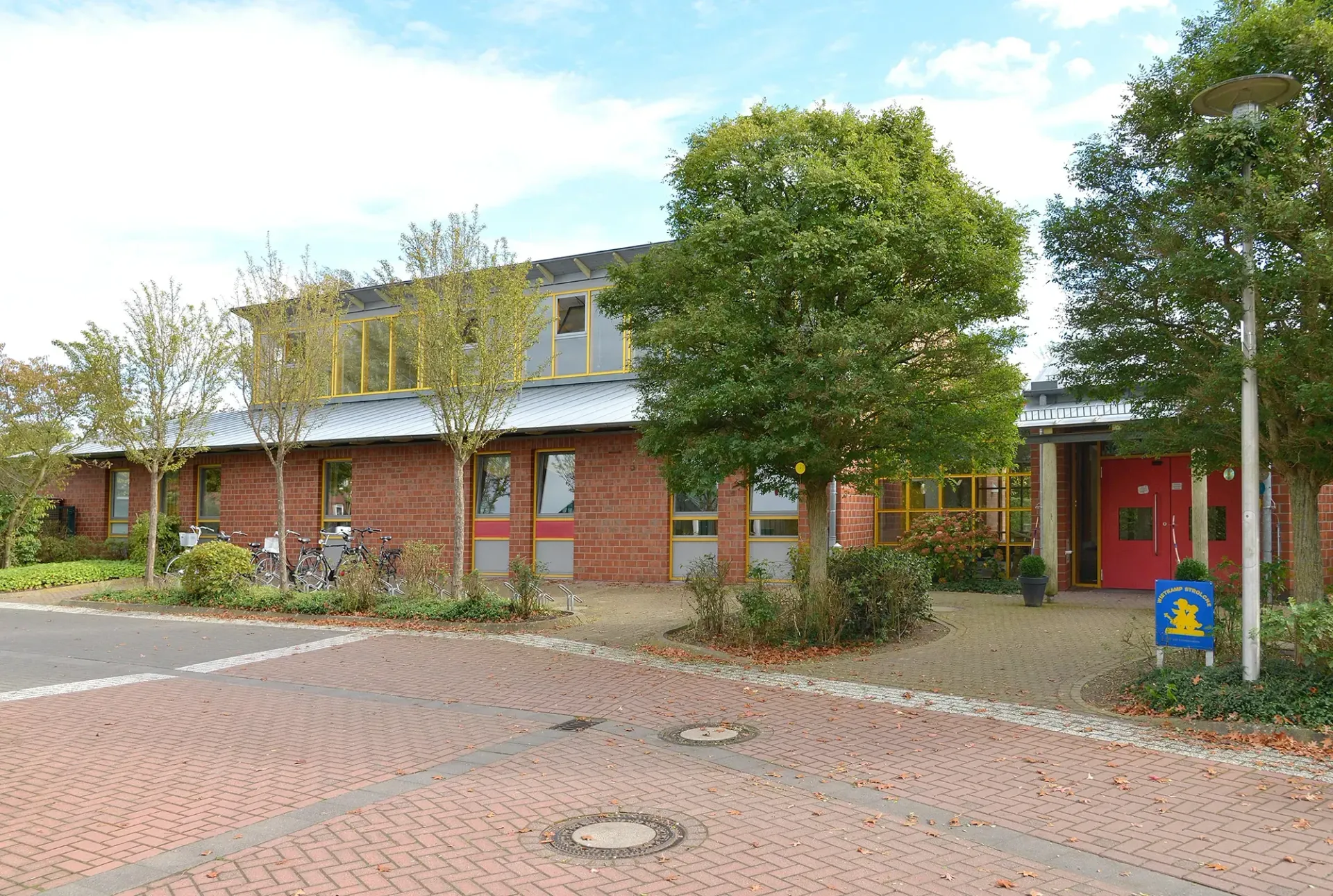 Ein Backsteingebäude mit einer roten Tür ist von Bäumen und einem Backsteinpflaster umgeben.