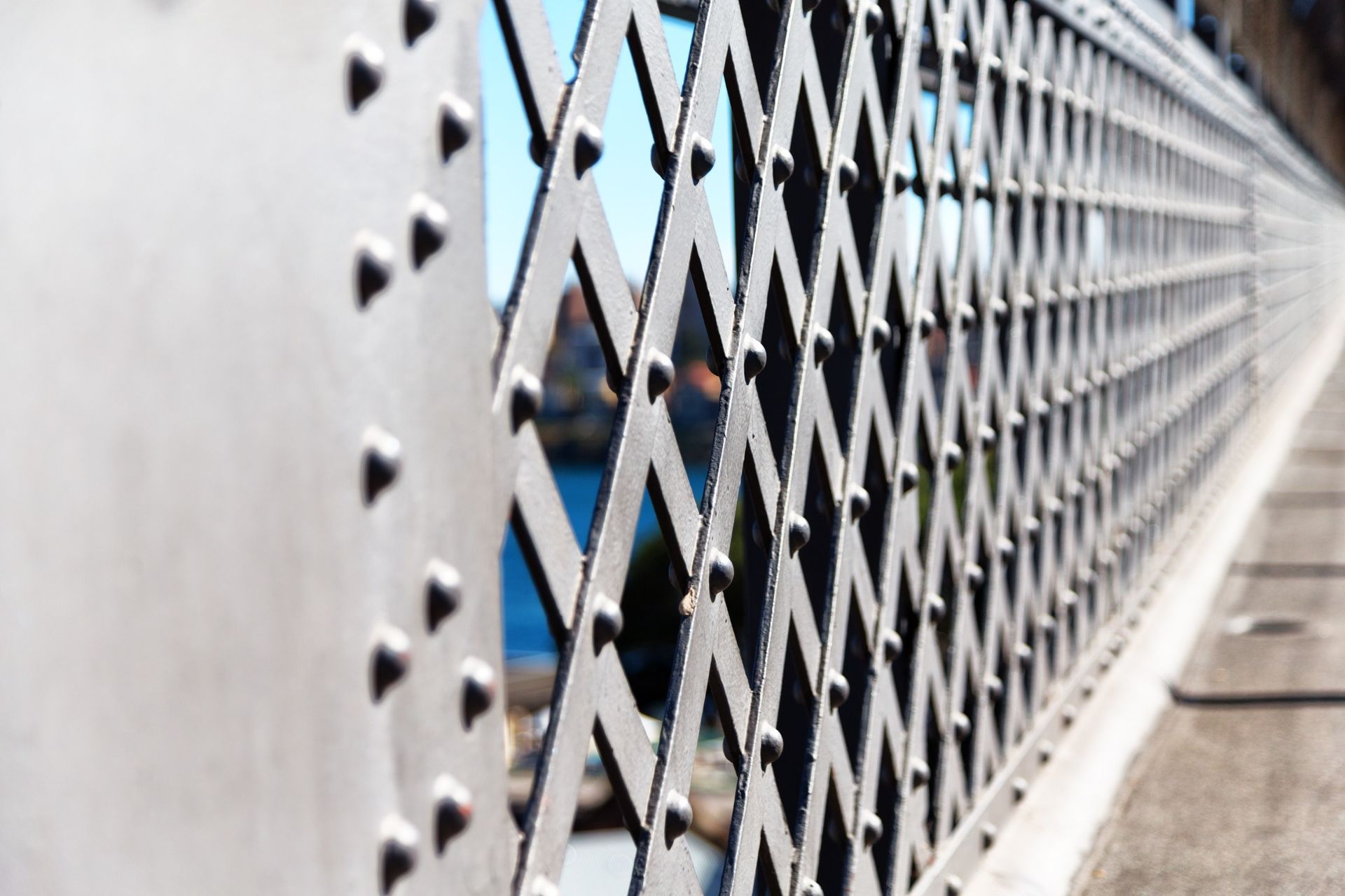 Primer plano de una barandilla de un puente de metal con un patrón de celosía de diamantes, remaches y un paisaje urbano borroso en el fondo.