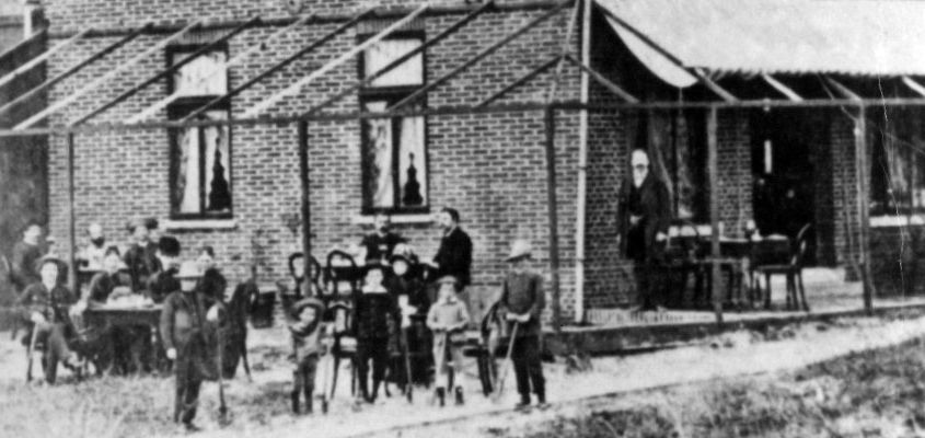 Eine Gruppe von Leuten steht vor einem Backsteingebäude. Schwarz-weiß-Aufnahme.