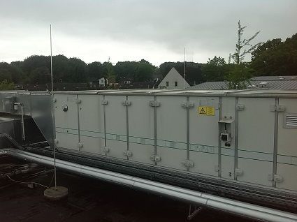 Eine große, industrielle Klimaanlage steht auf einem Flachdach vor dem Hintergrund von Bäumen und einem Gebäude.