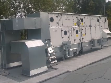 Auf einem grauen Dach steht eine große, industrielle Klimaanlage mit silbernen Metallkanälen.