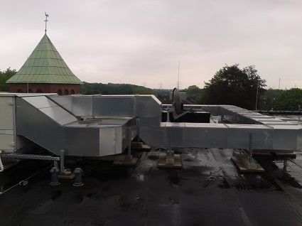 Auf dem Dach eines Flachbaus befinden sich metallene Lüftungskanäle; in der Ferne erhebt sich ein historischer Turm.