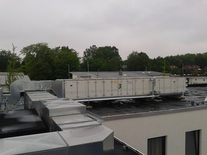Auf dem Dach eines Flachgebäudes steht eine große, industrielle Klimaanlage, die mit silbernen Lüftungskanälen verbunden ist.