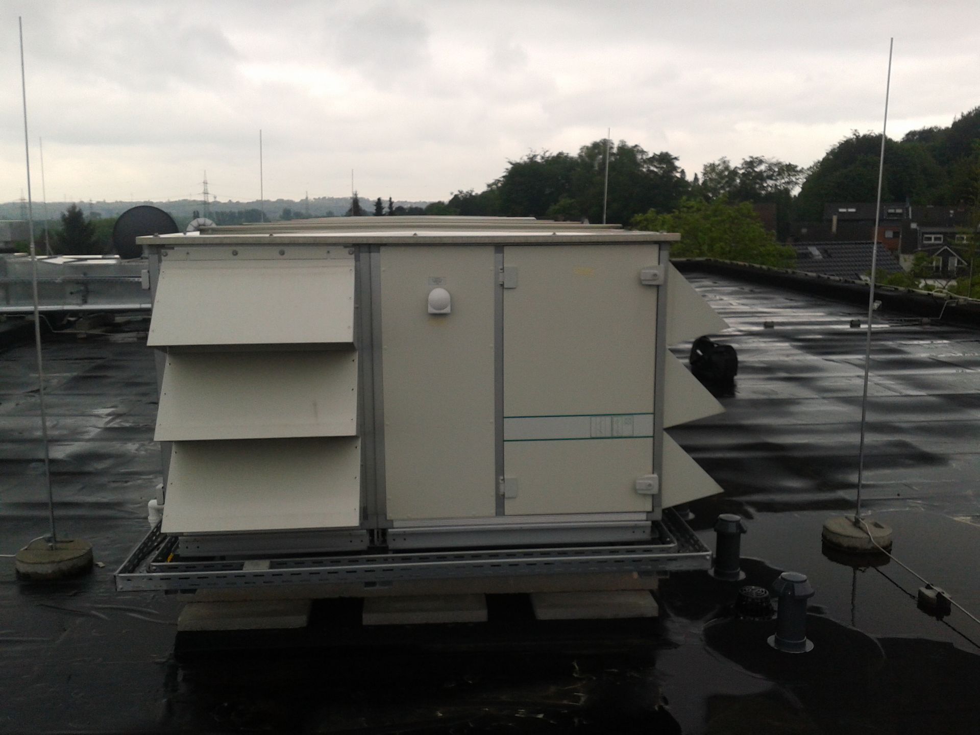 Eine große, helle, rechteckige Industrieklimaanlage steht auf einem nassen Flachdach unter bewölktem Himmel.