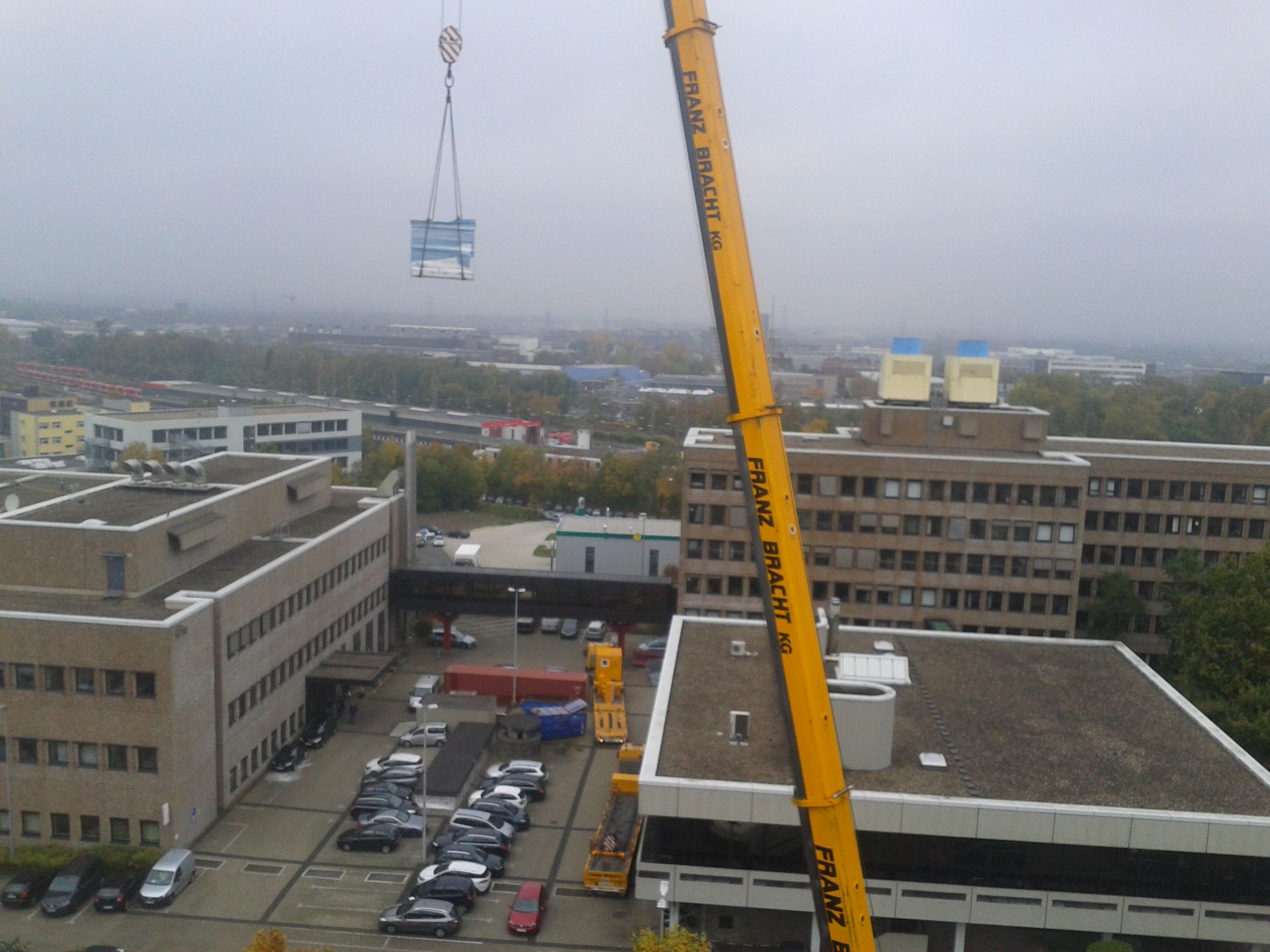 Ein gelber Mobilkran hebt eine große Klimaanlage über einen Bürogebäudekomplex mit einem darunterliegenden Parkplatz.