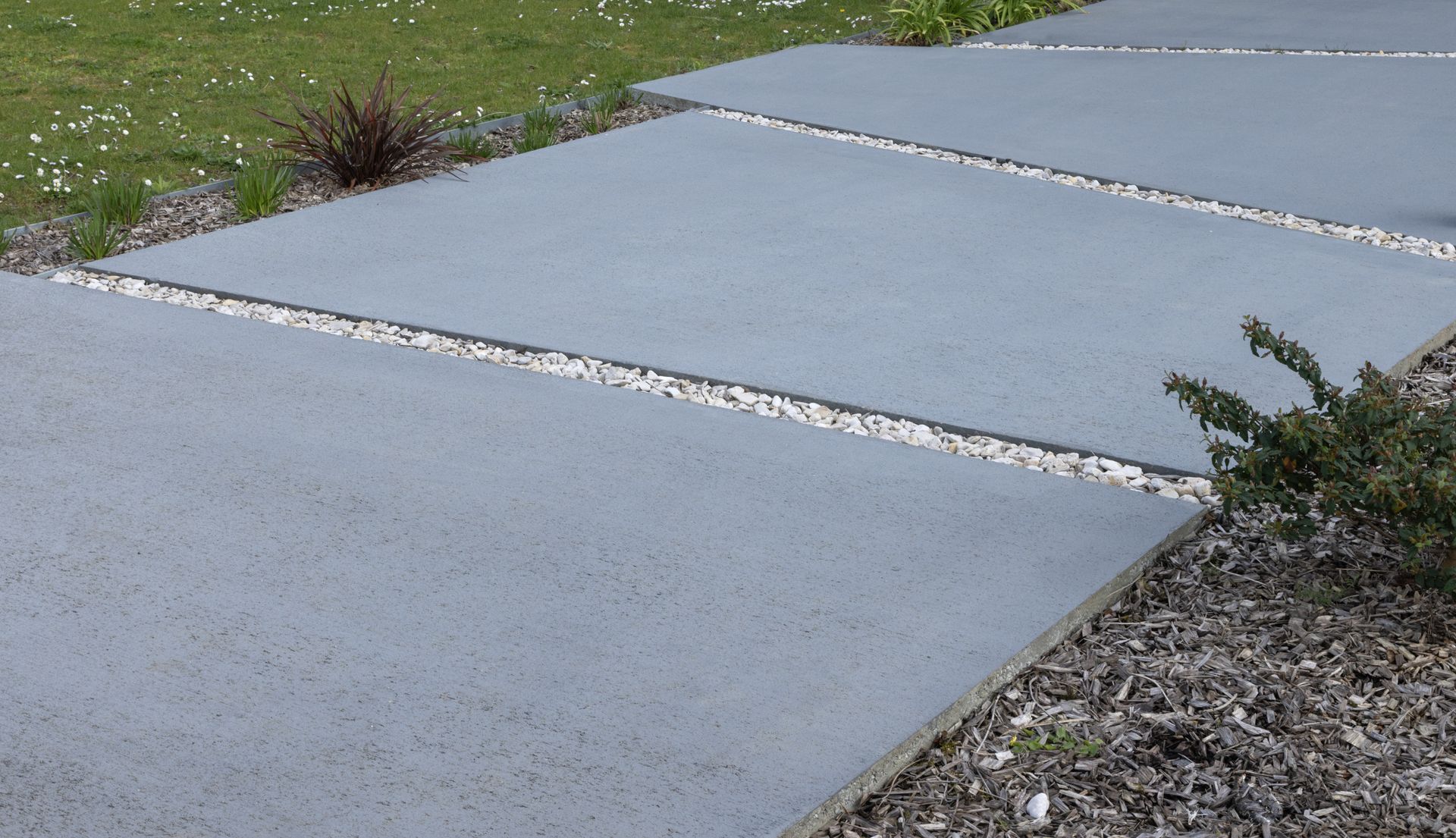 Dalles de trottoir en béton avec joints de gravier et petites plantes sur les bords.