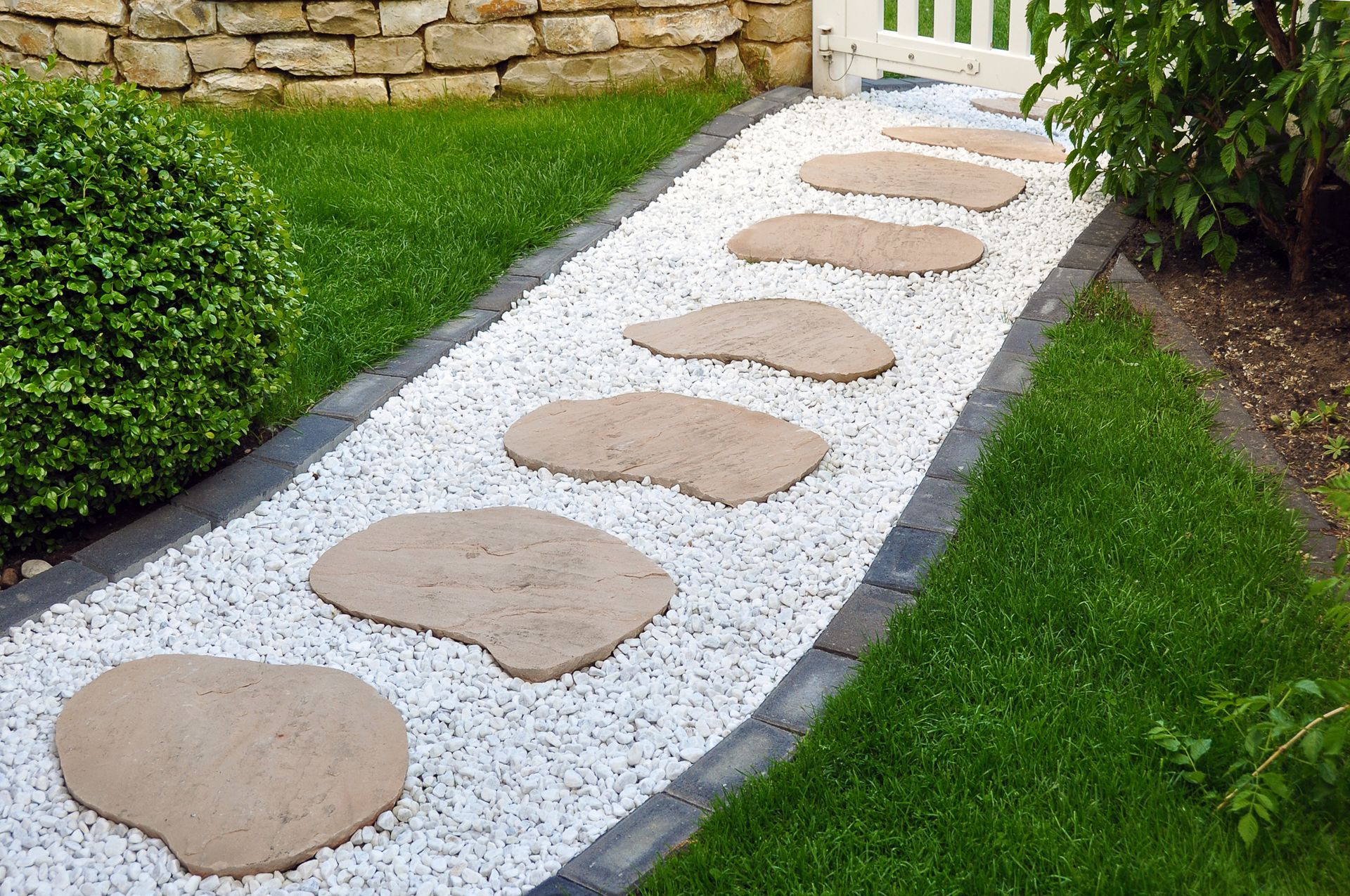 Allée de pierres traversant un jardin verdoyant, recouverte de gravier blanc, le long d'un mur de pierre.