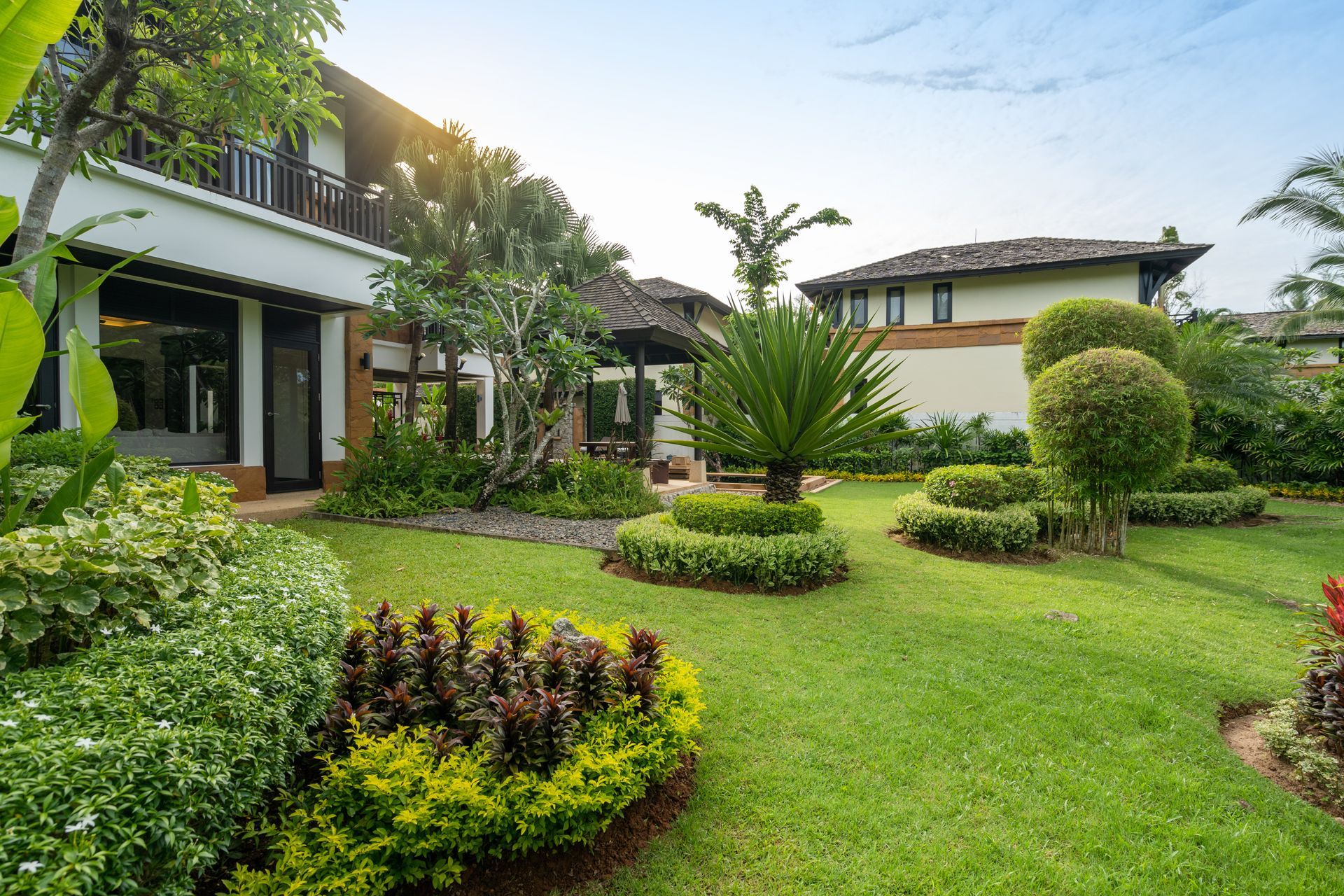 Jardin luxuriante avec pelouse soignée, arbustes et maisons modernes sous un ciel lumineux.