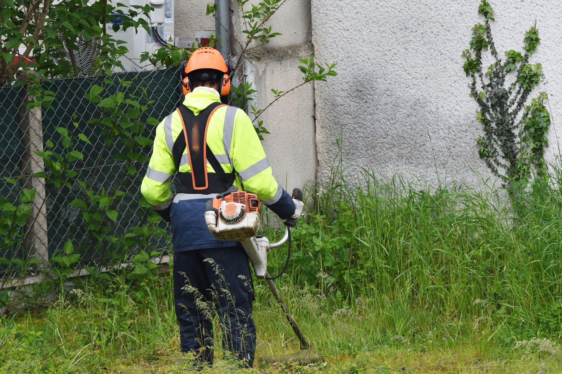 Un ouvrier en tenue réfléchissante utilise un coupe-bordures à côté d'une clôture souple et d'un mur en béton envahis par l'herbe.