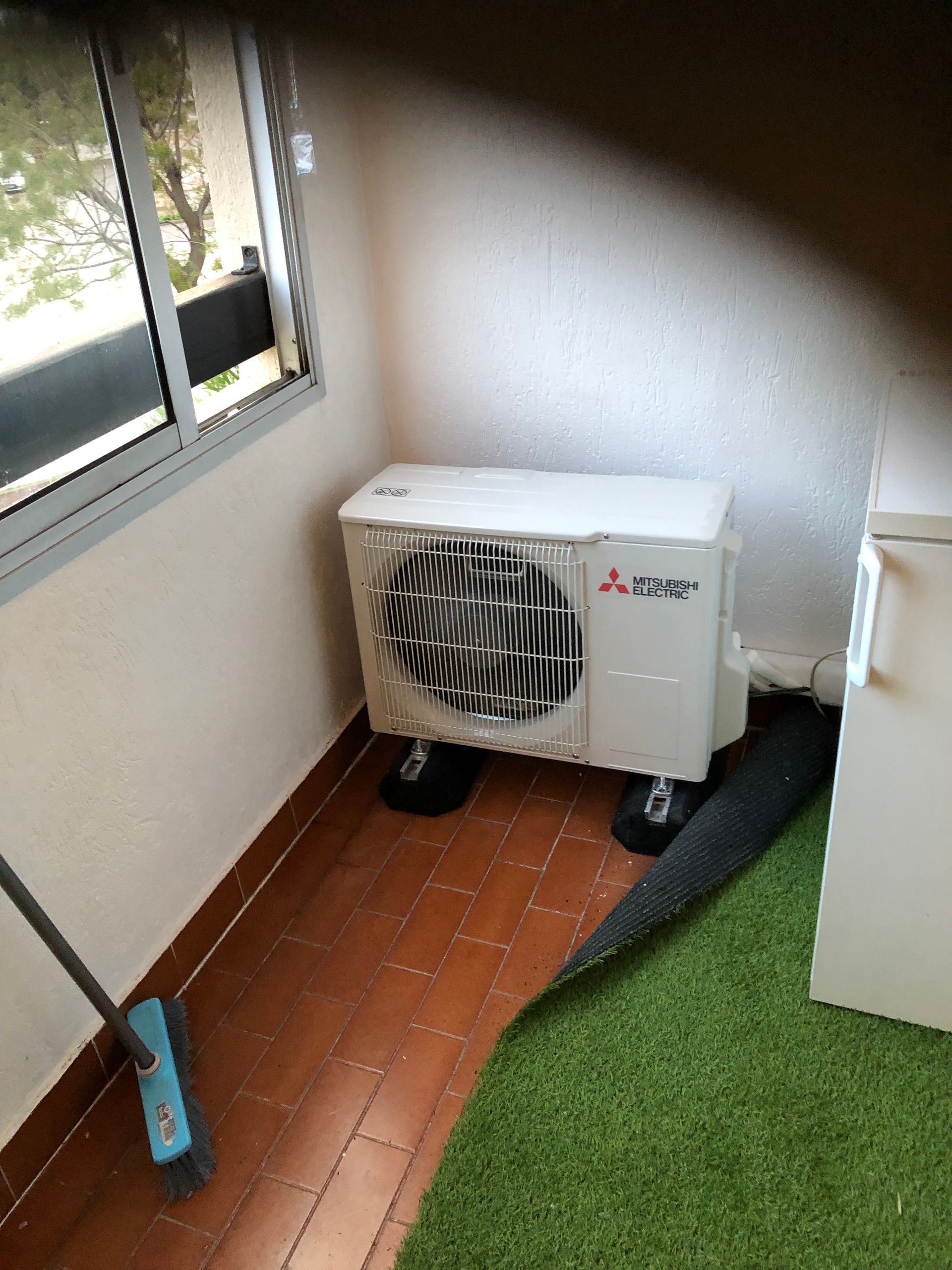 Petit coin de pièce avec un climatiseur sur roulettes à côté d'une fenêtre et un sol en gazon artificiel.