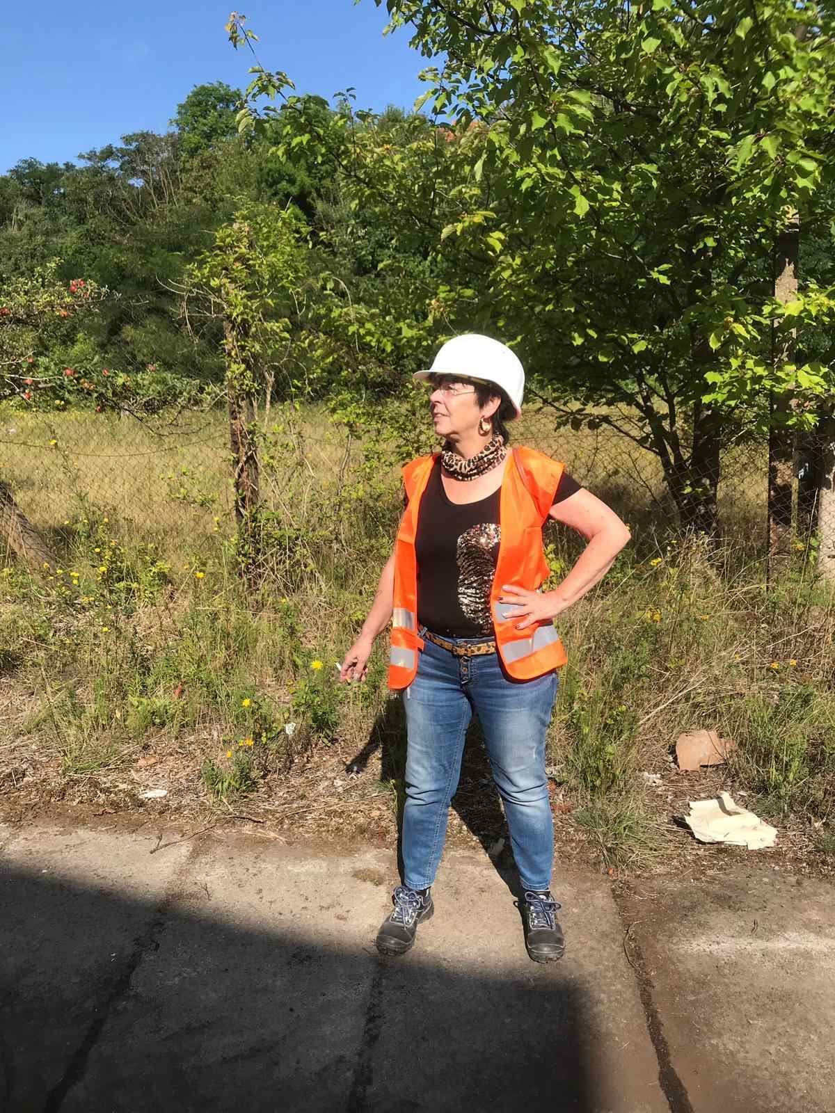 Eine Frau mit Schutzhelm und orangefarbener Weste steht auf einem Bürgersteig.