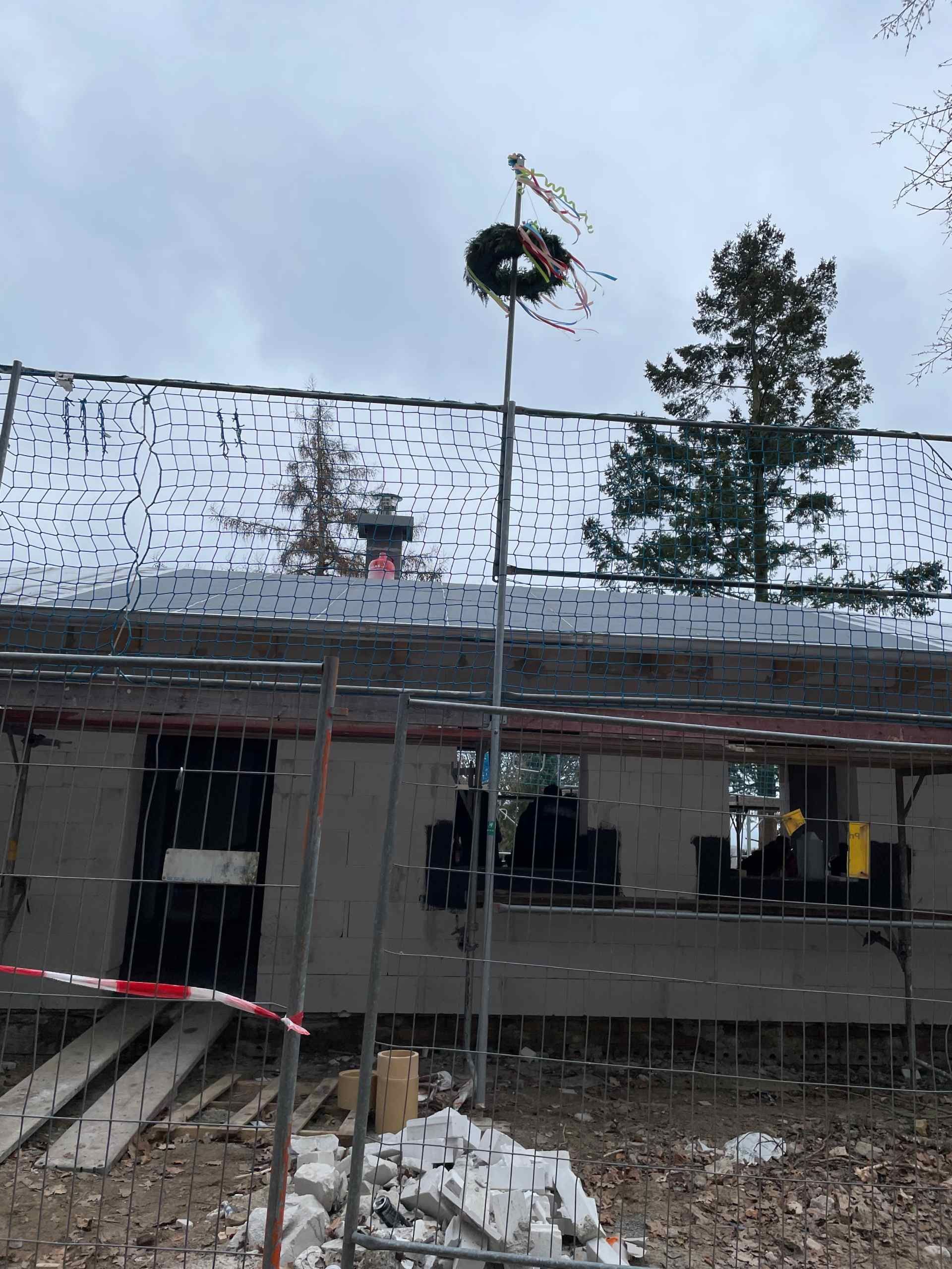 Vor einem im Bau befindlichen Gebäude hängt ein Kranz an einer Stange.