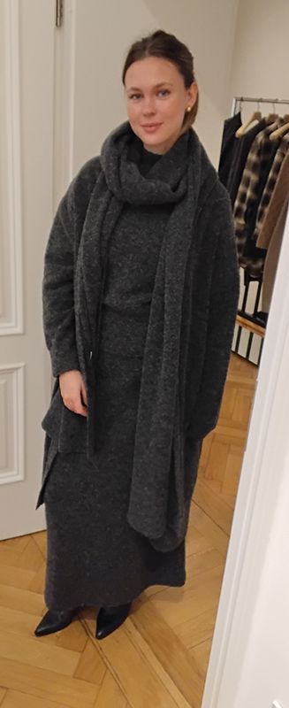 Frau in dunkelgrauem Mantel, Kleid und Schal steht im Innenbereich neben einer Wand und einem Türrahmen.
