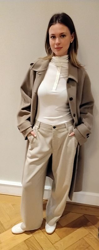Frau in hellbraunem Mantel, weißem Rollkragenpullover und cremefarbener Hose, Hände in den Taschen, posiert vor einer weißen Wand.