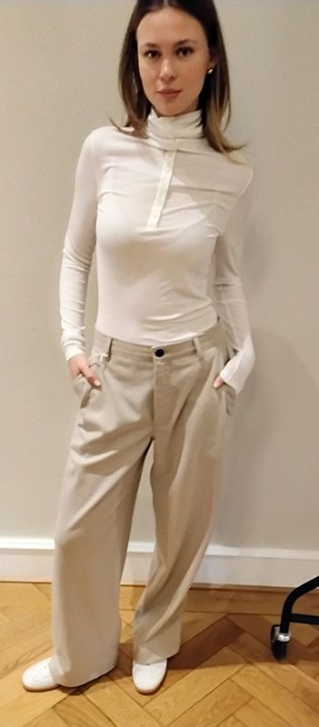 Frau in weißem Rollkragenoberteil und beiger Hose mit weitem Bein, steht mit den Händen in den Taschen.