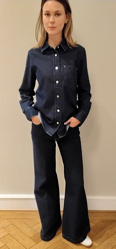 Frau in dunklem Jeanshemd und Schlagjeans, die Hände in den Taschen. Stehend vor einer beigen Wand.