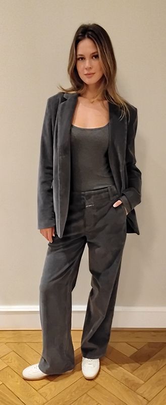 Frau in grauem Anzug und weißen Schuhen steht drinnen auf einem Holzboden.