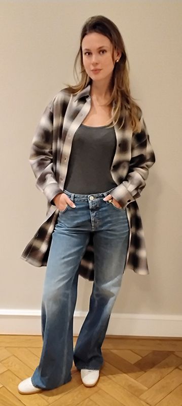 Frau in kariertem Hemd, grauem Top und Schlagjeans, Hände in den Taschen, an eine Wand gelehnt.