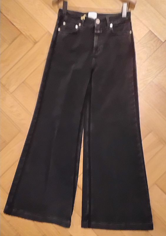 Schwarze, verkürzte Jeans mit weitem Bein, die an einem hölzernen Kleiderbügel hängt.