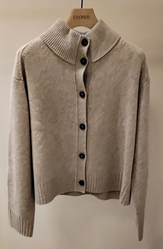 Beige Strickjacke mit hohem Kragen, Knopfleiste und langen Ärmeln hängt an einer Kleiderstange.