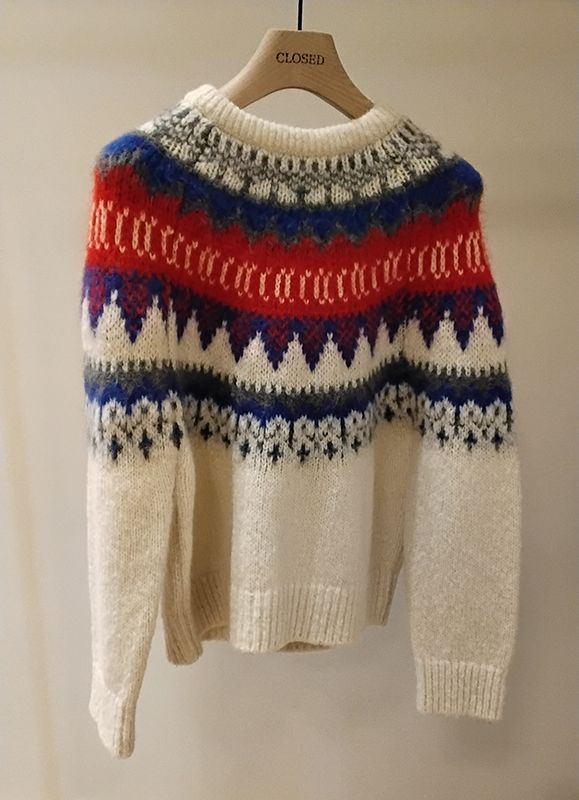 Weißer und gemusterter Pullover mit roten, blauen und grauen Akzenten hängt auf einem hölzernen Kleiderbügel.