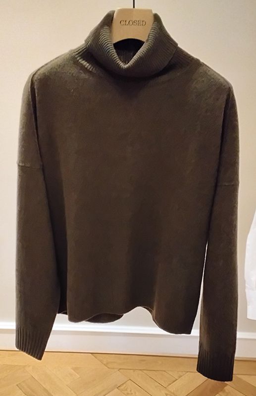 Brauner Rollkragenpullover auf einem hölzernen Kleiderbügel vor einer beigen Wand.