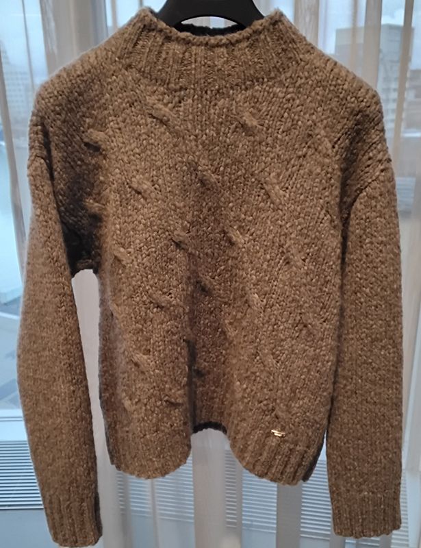 Brauner Zopfmusterpullover mit hohem Kragen, hängt auf einem schwarzen Kleiderbügel.