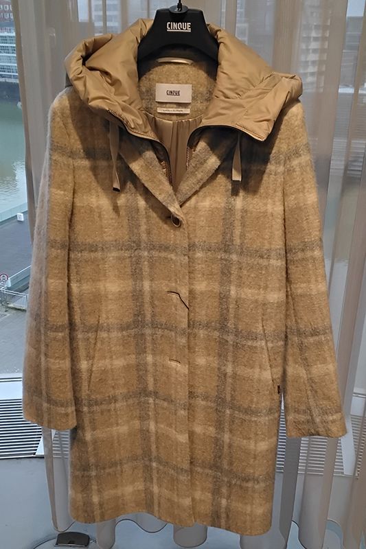 Beiger karierter Wollmantel mit hellbrauner Kapuze hängt auf einem schwarzen Kleiderbügel vor einem Fenster.