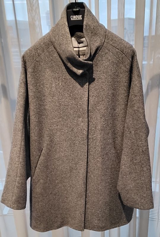 Grauer Wollmantel mit breitem Kragen, der auf einem Kleiderbügel vor einem Fenster hängt.