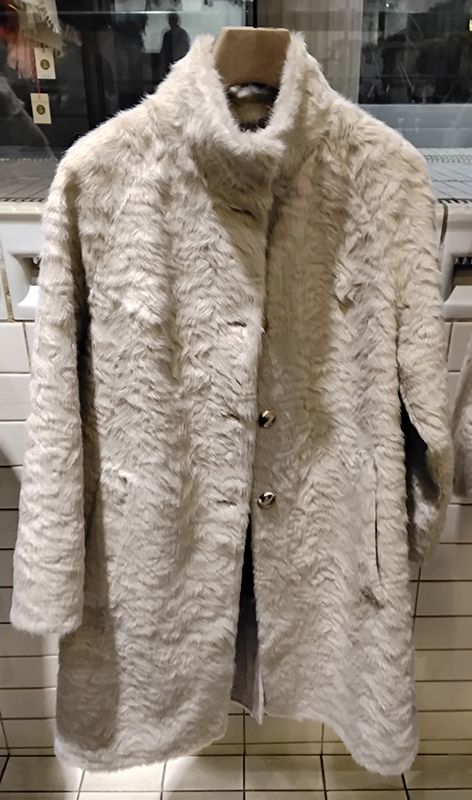 Ein hellfarbiger, strukturierter Kunstpelzmantel mit hohem Kragen hängt an einem Kleiderständer.