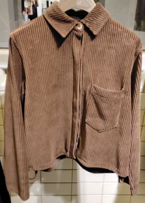Braunes Cordhemd mit einer einzelnen Brusttasche, Knopfleiste und Kragen, das an einem Kleiderständer hängt.