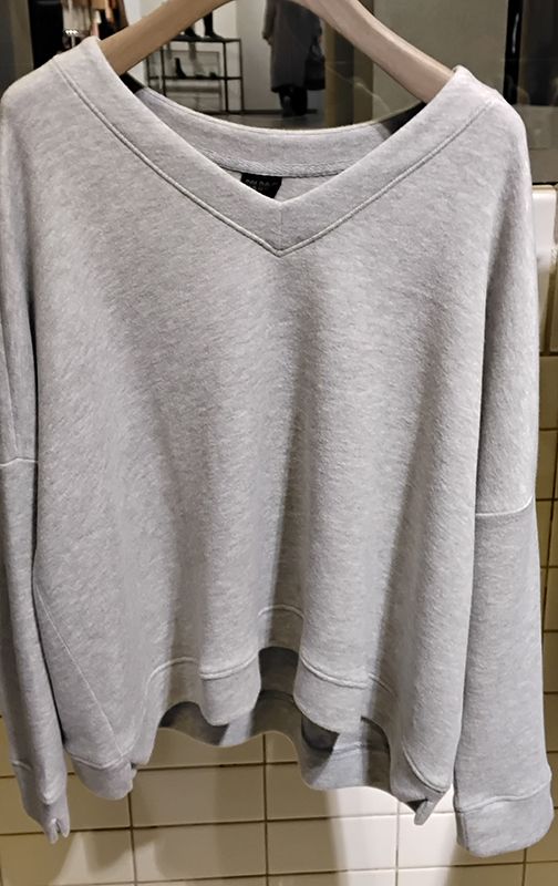 Graues Sweatshirt mit V-Ausschnitt hängt an einem Holzkleiderbügel; im Hintergrund sind Fliesen zu sehen.