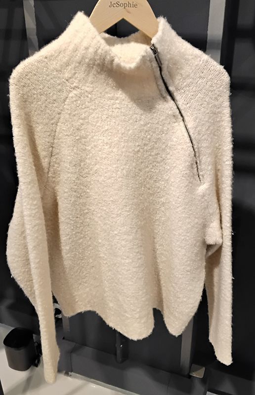 Cremefarbener, flauschiger Pullover mit schwarzem diagonalen Reißverschluss, hängt an einer Stange.