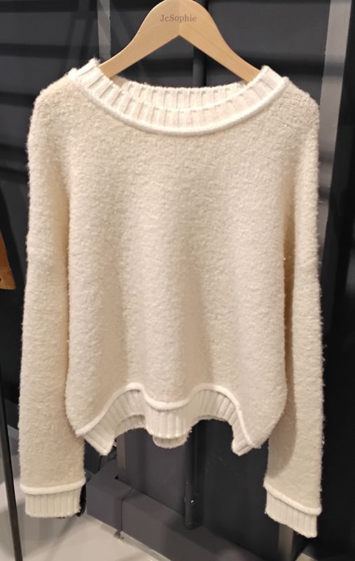 Cremefarbener strukturierter Pullover auf einem Holzbügel mit weißem Besatz um Hals, Bündchen und Saum.