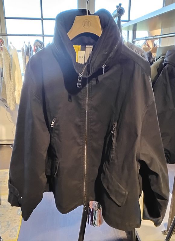 Schwarze Jacke mit großem Kragen und asymmetrischem Reißverschluss, die an einem Kleiderständer hängt.