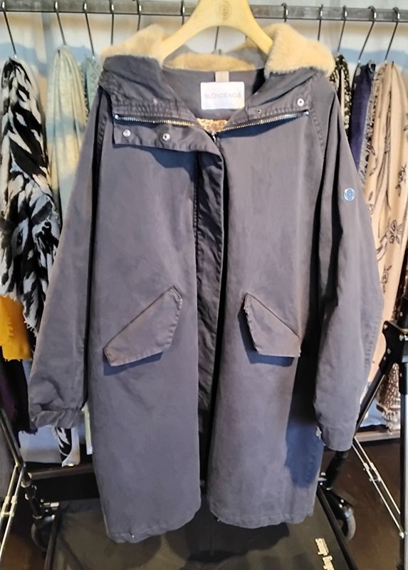 Grauer Parka mit Kapuze und Fellfutter, hängt zusammen mit anderer Kleidung an einem Kleiderständer.