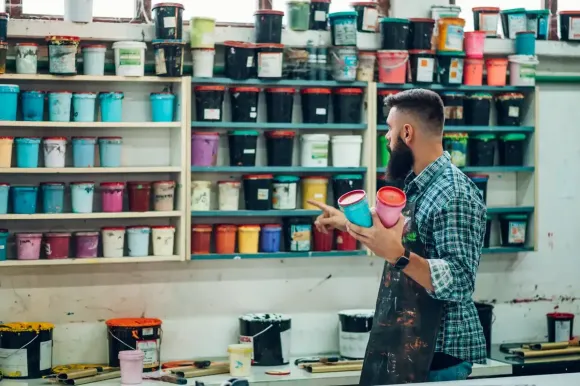 Hombre con barba en el taller, sosteniendo baldes de pintura, apuntando hacia una pared de latas de pintura de colores.