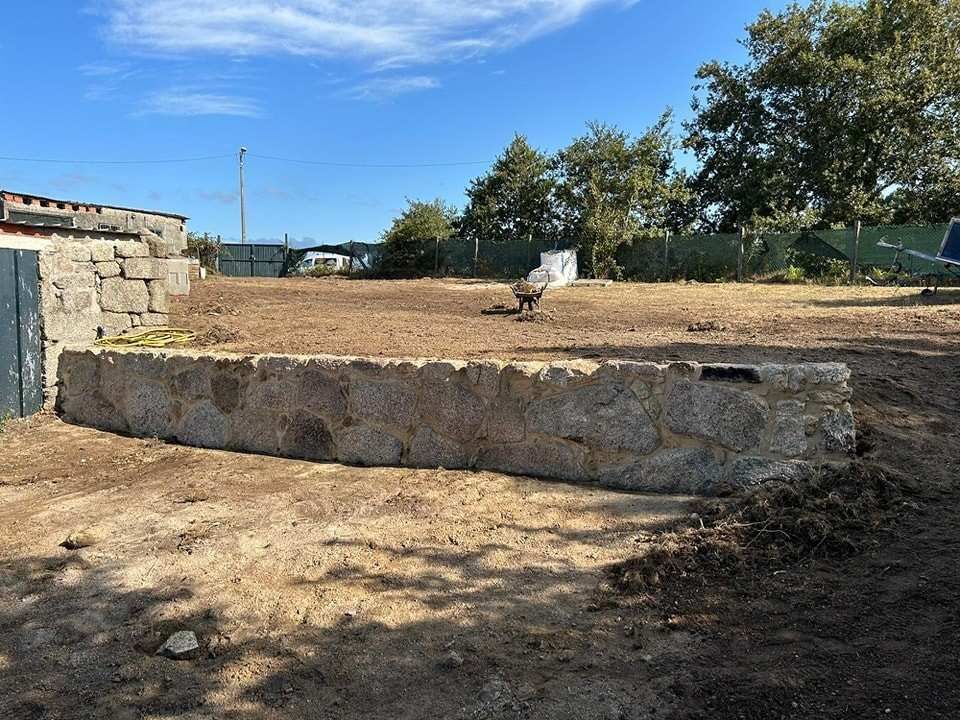 Un muro bajo de piedra se alza en un patio cubierto de tierra en un día soleado, con árboles y una valla al fondo.