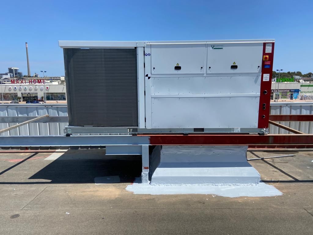 Unidad de aire acondicionado en azotea, de color gris claro y rojo, en un edificio comercial con cielo azul.