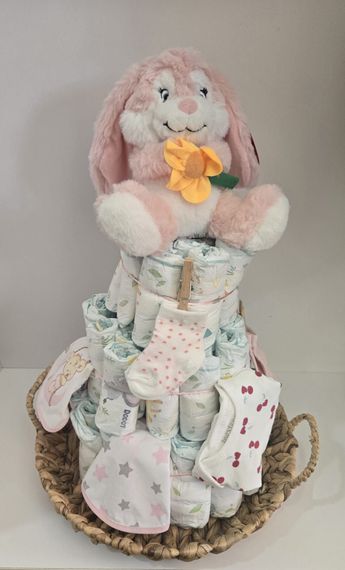 Torta de pañales con adorno de conejito rosa, calcetines, baberos y toallitas en una canasta tejida.