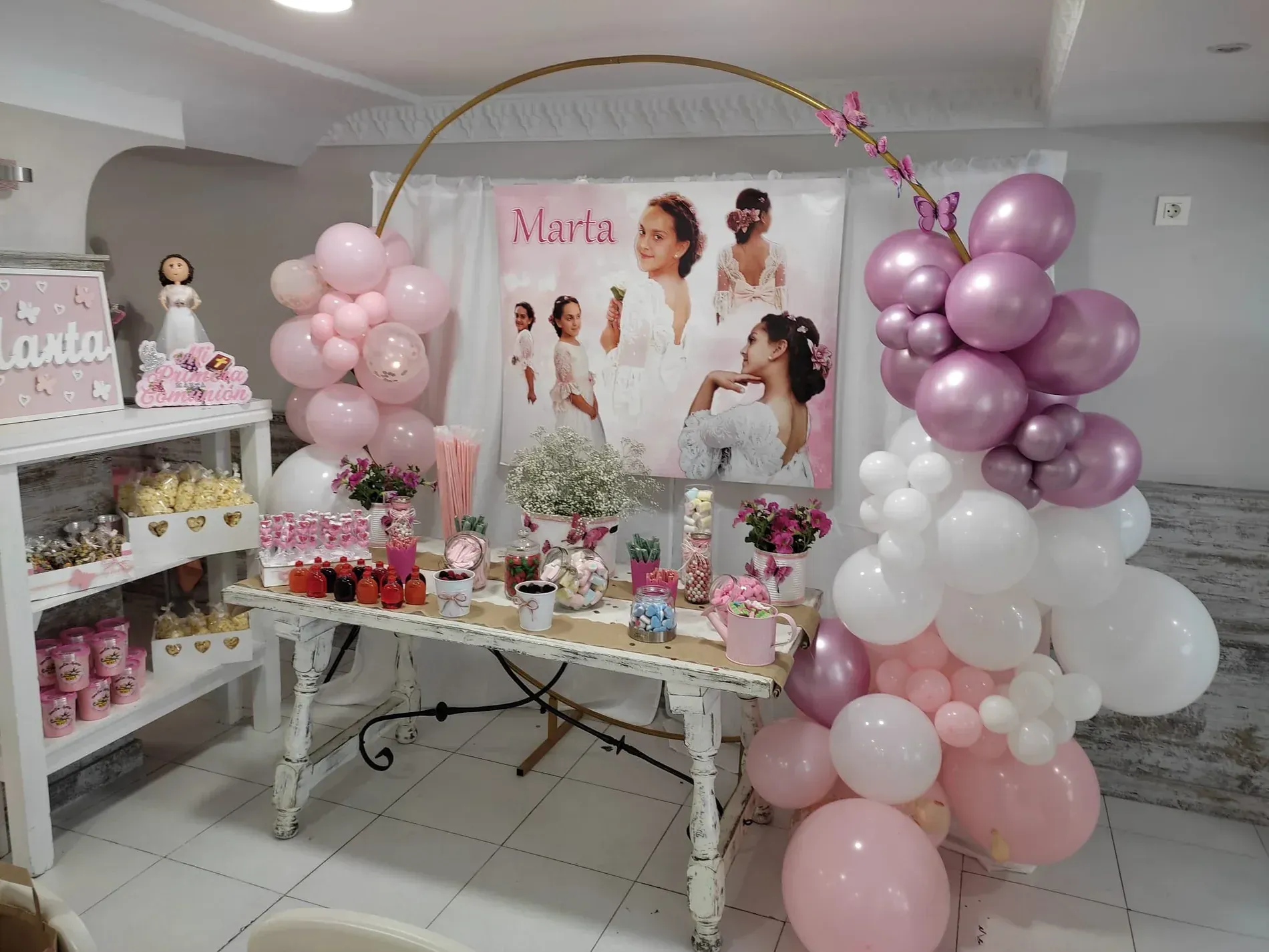Decoración de fiesta con globos, fondo con una niña, mesa con golosinas, combinación de colores rosa y blanco.