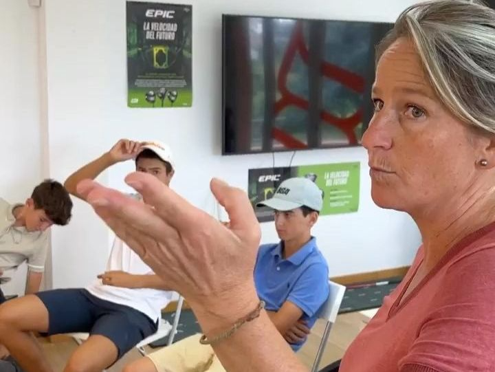 Mujer haciendo gestos, hablando con un grupo de hombres jóvenes en una habitación, con carteles en la pared, en un entorno informal.