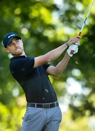 Golfista con camisa negra, pantalón gris y gorra balancea su palo de golf en un campo verde, al aire libre.
