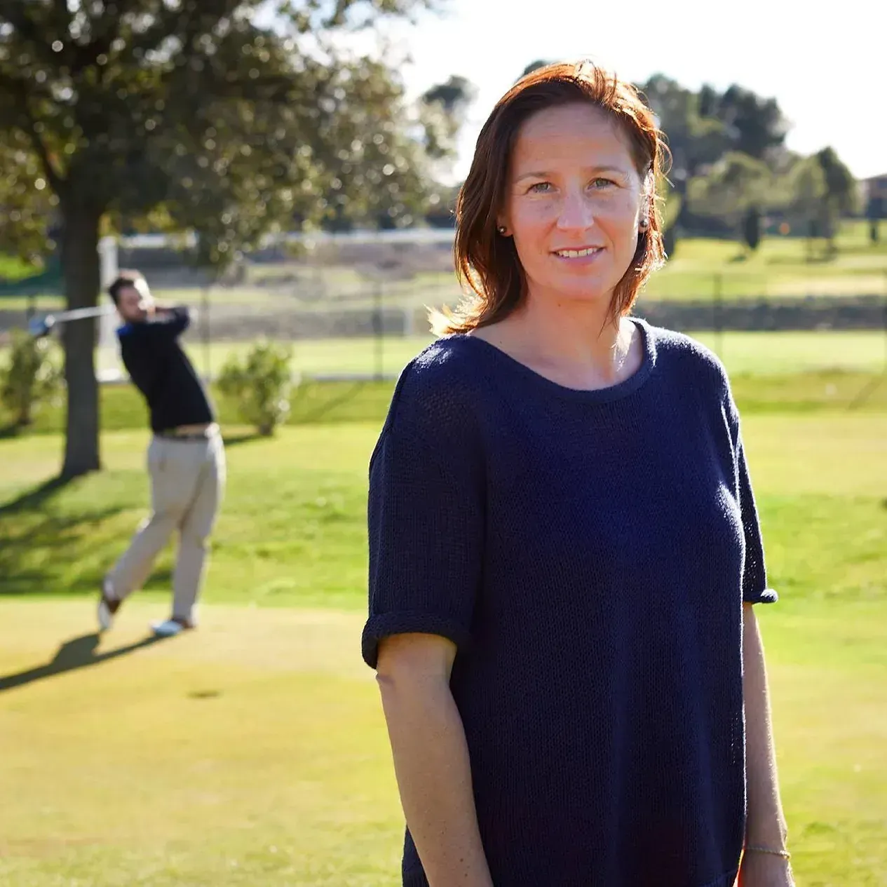 Mujer sonriendo en un campo de golf soleado, el hombre en el fondo balancea un palo de golf.