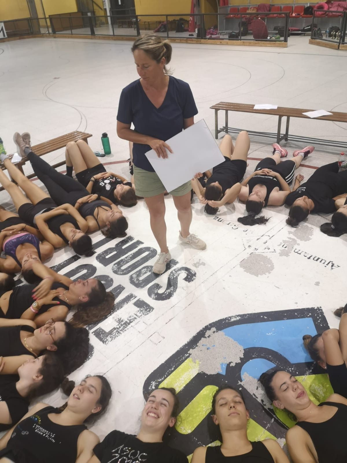 Una mujer da instrucciones a un grupo de jóvenes tumbadas en el suelo de un gimnasio. Vestían de negro y algunas tenían los ojos cerrados.