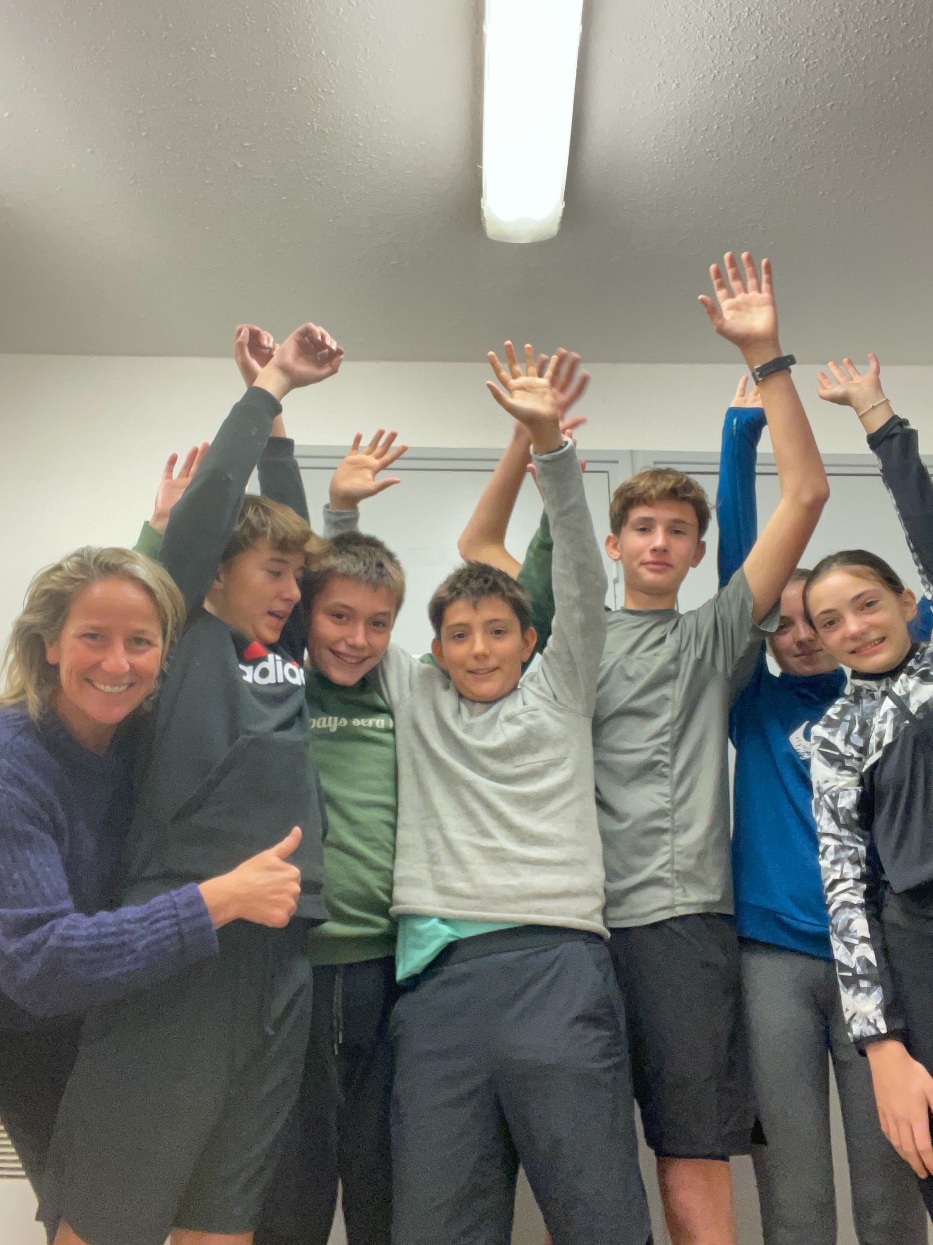 Un grupo de estudiantes sonrientes y un profesor con los brazos levantados, en una habitación muy iluminada.